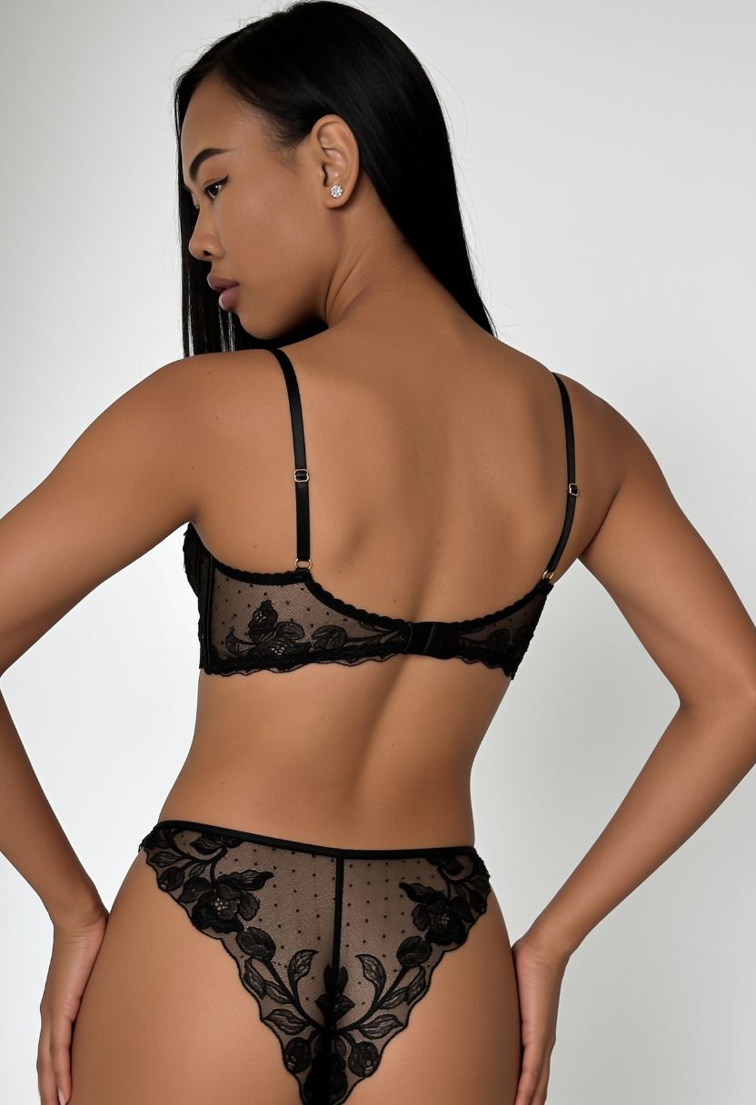 A woman shown from the back wearing a black lace lingerie set with floral lace trim and sheer mesh panels, photographed against a light gray studio background.